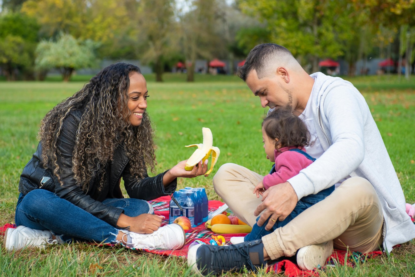 Two parents sat with child on the grass. The mother is feeding the child a banana,